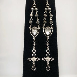 May include: Silver earrings with a black bead chain, a silver heart with a crown and a silver cross with a black background.