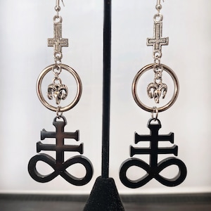 May include: A pair of black and silver earrings featuring a black upside-down cross with an infinity symbol, a silver circle with a ram's head charm, and a silver upside-down cross.