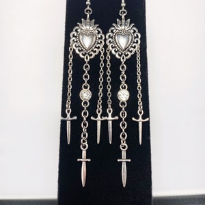 May include: Silver dangle earrings with a heart-shaped charm, a crystal, and multiple sword charms hanging from a chain.