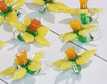 以下が含まれることがあります： 手作りのガラス製水仙の花のオーナメント。各花は黄色の花びら、オレンジ色のラッパ、緑色の茎が特徴です。オーナメントには、吊り下げ用の銀色のリングが付いています。水仙は白い表面に配置されています。