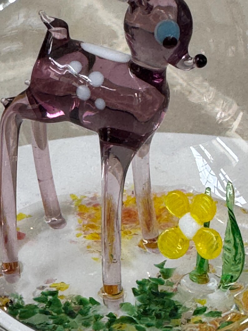 以下が含まれることがあります： 透明感のある紫色のガラス製シカの置物。透明な台座の上に置かれ、周囲には黄色と緑色のガラスの破片が散らばっています。シカの背中には白い斑点があり、黒い鼻と青い目をしています。そばには白い中心部を持つ小さな黄色いガラスの花が添えられています。