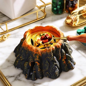 May include: A volcano-shaped wax warmer with a gold-colored interior and a black and orange exterior. A small gold-colored ladle holds red wax cubes. The warmer is designed for melting wax for various crafts and projects.