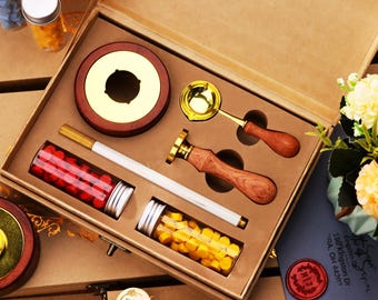 May include: A wax seal kit in a brown box. The kit includes a wooden and gold wax warmer, a gold-colored wax spoon, a wax stamp with a wooden handle, two glass jars filled with red and yellow wax beads, and several wax seal stamps.