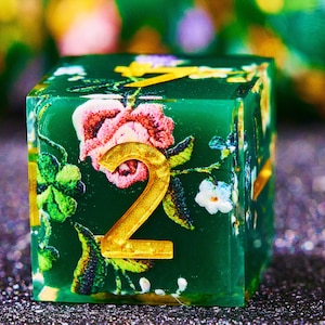 May include: A handcrafted, square, green resin die with floral patterns and gold-colored numerals. The die features a vibrant design of pink, white, and blue flowers with green leaves. The number "2" is prominently displayed in gold.