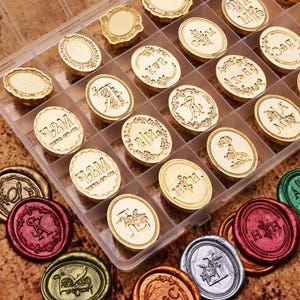 Könnte beinhalten: Eine Sammlung goldfarbener Wachssiegelstempel mit verschiedenen Designs, darunter Initialen, Blumenmuster und dekorative Bordüren. Die Stempel sind in einem transparenten Kunststoff-Organizer angeordnet, darunter sind farbige Wachssiegel zu sehen.