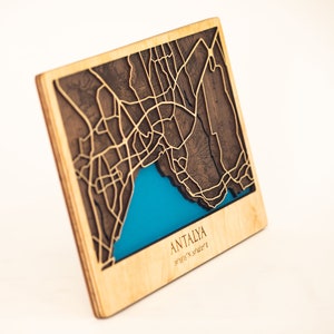 May include: A wooden map of Antalya, Turkey, with a blue inlay representing the Mediterranean Sea. The map is laser-cut and features detailed street outlines. The city name and coordinates are engraved below the map.