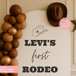 Może przedstawiać: Ozdobny znak z napisem "Levi's first Rodeo" w kolorze czarnym. Znak jest ozdobiony ilustracją kapelusza kowbojskiego, brązowym kapeluszem kowbojskim, gwiazdą i łukiem balonowym w odcieniach brązu i złota.