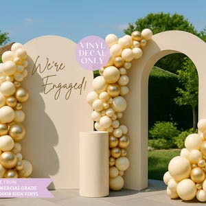 May include: An engagement photo backdrop with a cream-colored arch and a cylindrical pedestal. The backdrop is decorated with cream and gold balloons. The words "We're Engaged!" are written on the backdrop.