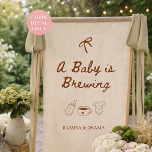 May include: A beige fabric sign with the words "A Baby is Brewing" in brown script. The sign features illustrations of a baby bottle, a cup of coffee, and a onesie. The names "RAMISA & OSAMA" are printed at the bottom.