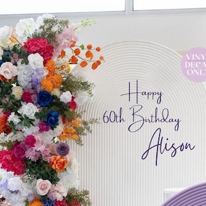 May include: A vibrant floral display with red, white, blue, and orange flowers on the left of a white, arched backdrop. The backdrop has the text "Happy 60th Birthday Alison" in dark purple script, suitable for a 60th birthday celebration.
