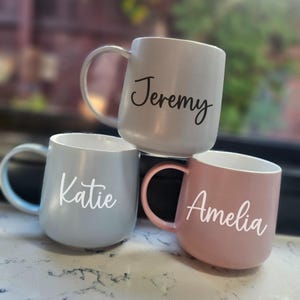 May include: Three ceramic mugs with personalised names. The mugs are in shades of grey, blue, and pink. The names "Jeremy", "Katie", and "Amelia" are written in white on the mugs.