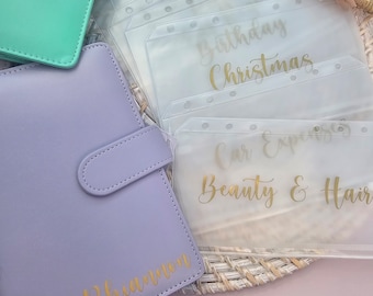 May include: A set of clear plastic pockets with gold lettering for a personal organizer. The pockets are labeled "Birthday", "Christmas", "Car Expenses", and "Beauty & Hair". A purple leather planner with the name "Rhiannon" is in the foreground.