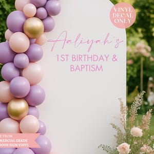 May include: A white arch-shaped sign with pink text reading "Aaliyah's 1st Birthday & Baptism". A balloon garland in pink, purple, and gold decorates the left side. The sign is made from outdoor sign vinyl.