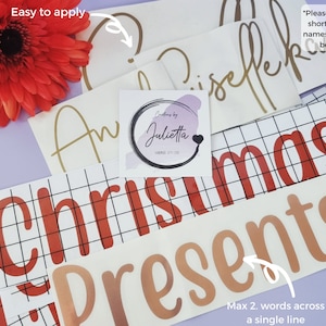 May include: A collection of adhesive vinyl decals featuring various names and the words Christmas Presents in red and metallic gold lettering. The decals are displayed on transfer paper with grid lines, accompanied by a square logo sticker.