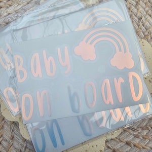 May include: A set of four car decals with a rose gold finish. The decals read "Baby on board" and feature a rainbow with clouds.