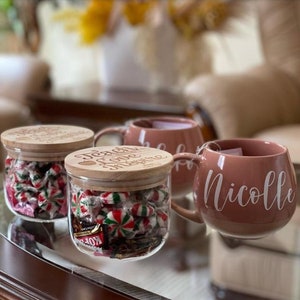 May include: Two clear glass jars with wooden lids engraved with the text Teach Love Inspire, filled with red, white, and green candies, alongside two dusty rose ceramic mugs with the white script name Nicolle printed on the side.
