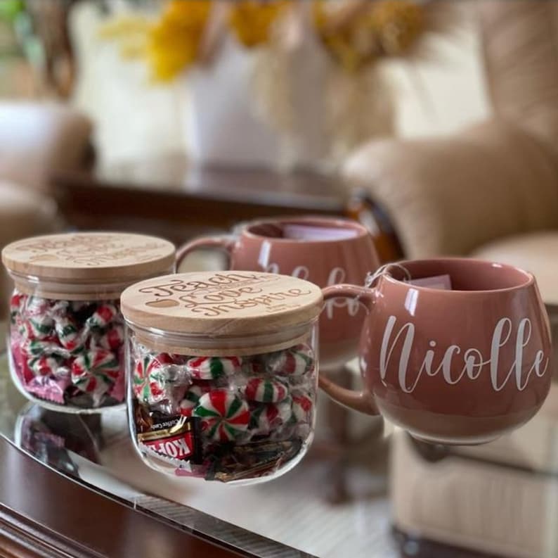 May include: Two clear glass jars with wooden lids engraved with the text Teach Love Inspire, filled with red, white, and green candies, alongside two dusty rose ceramic mugs with the white script name Nicolle printed on the side.