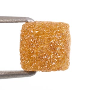 May include: A rough, square-cut, golden-brown diamond held by a silver tweezer. The diamond has a textured surface with many facets, reflecting light. The gemstone is set against a white background.