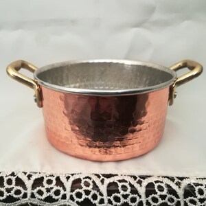 Peut inclure: Petite casserole en cuivre avec une texture martelée et des poignées en laiton. L'intérieur est argenté. Le pot est posé sur une surface blanche avec une bordure en dentelle décorative. Idéal pour la cuisine et le service.