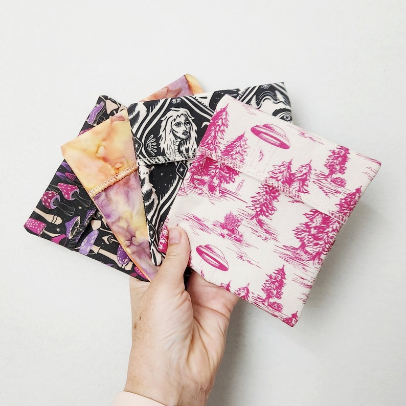 May include: Four reusable food wraps with different designs. One wrap features a black and white illustration of a woman with a crown, another has a pink and white floral pattern, another has a pink and white pattern with a UFO, and the last one has a black and white mushroom pattern.