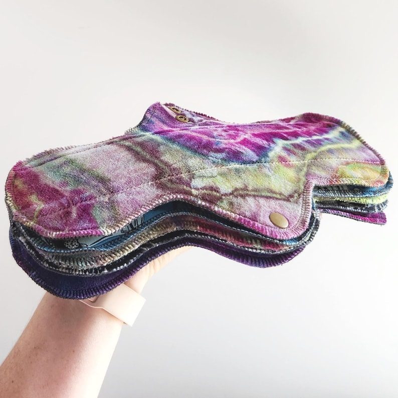 May include: A stack of reusable cloth menstrual pads featuring a vibrant tie-dye pattern in shades of magenta, purple, and green. The pads have a soft, textured surface, contoured edges, and are secured with small metallic snap buttons on the wings.