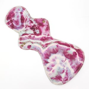 May include: A reusable cloth menstrual pad featuring a tie-dye pattern in shades of magenta, deep purple, and white. The pad has an hourglass shape with stitched edges and two small metallic snap fasteners on the side wings for secure closure.