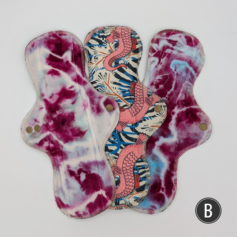 May include: Three reusable cloth pads with contoured shapes and metal snap closures. Two pads feature a deep magenta and white tie-dye pattern, while the center pad displays a pink lizard print on a cream background with blue foliage accents.
