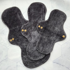 May include: Three charcoal gray reusable cloth menstrual pads with gold snaps. The pads are layered on top of each other.