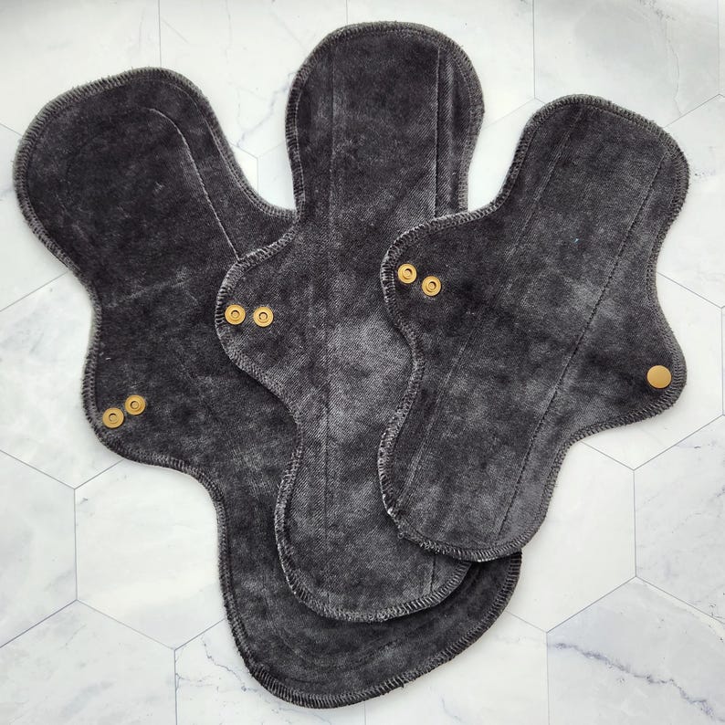May include: Three charcoal gray reusable cloth menstrual pads with gold snaps. The pads are layered on top of each other.