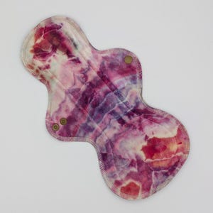 May include: A reusable cloth menstrual pad featuring an abstract tie-dye pattern in shades of magenta, purple, and white. The hourglass-shaped pad has stitched edges and two small metallic snap fasteners on the side wings for secure closure.