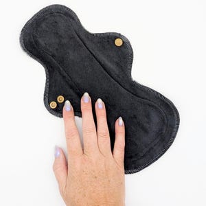 May include: A black reusable cloth menstrual pad with a contoured hourglass shape and dark grey stitched edges. The pad features three metallic gold-colored snap fasteners on the wings, designed to secure the item around undergarments.