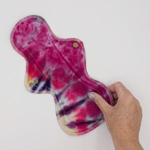 May include: A reusable cloth menstrual pad featuring a vibrant tie-dye pattern in shades of magenta, purple, and yellow. The pad has a contoured hourglass shape with stitched edges and two small metallic snap fasteners on the side wings for closure.