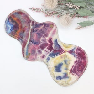 May include: A reusable cloth menstrual pad with a tie-dye pattern in shades of purple, blue, yellow, and orange. The pad has a curved shape with snap closures and a stitched edge.