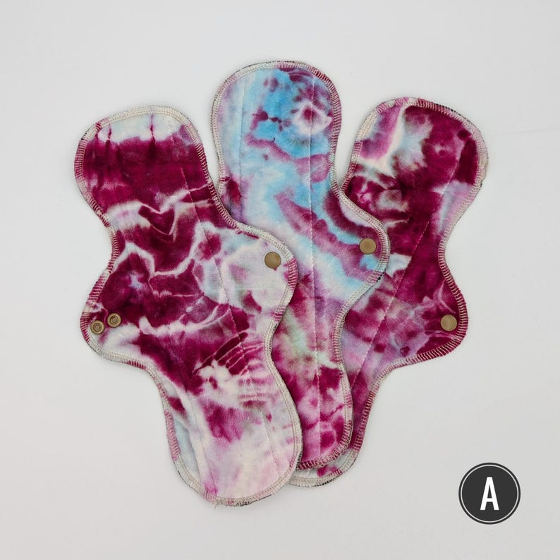 May include: Three reusable cloth menstrual pads featuring a tie-dye pattern in shades of deep magenta, white, and light blue. Each pad has a contoured shape with metal snap fasteners on the side wings to secure the fabric in place during use.