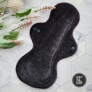 May include: A dark gray reusable cloth menstrual pad with a 12-inch flare. The pad has two black snaps on the front and one on the back.