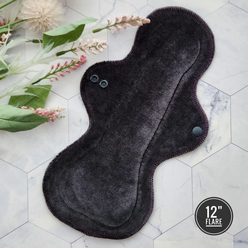 May include: A dark gray reusable cloth menstrual pad with a 12-inch flare. The pad has two black snaps on the front and one on the back.