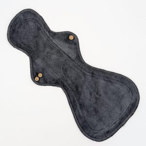 May include: A charcoal gray reusable cloth menstrual pad with an hourglass shape and a soft, textured fabric surface. The pad features dark stitching along the edges and three small, circular metallic snap fasteners located on the side wings.