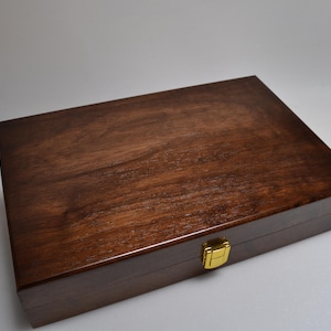 Può includere: Una scatola di legno rettangolare con finitura marrone scuro e chiusura dorata. La scatola ha una superficie liscia e lucida e la venatura del legno è visibile. La scatola è chiusa e poggia su una superficie bianca.