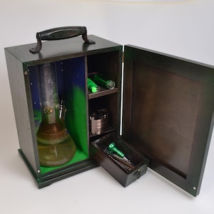 May include: A wooden box with a hinged lid, containing a glass bong, a grinder, and other smoking accessories. The box is painted dark green and has a black handle.