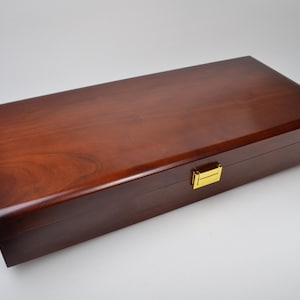 May include: A rectangular wooden storage box with a smooth, polished dark brown finish and visible wood grain. The box features a hinged lid and a single, centered gold-colored metal latch on the front edge for secure closure.