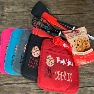 May include: Four red, blue, gray, and pink oven mitts with the text "One Smart Cookie" and a cookie with glasses on it. A red spatula, a whisk, a red silicone brush, a red measuring spoon, and a bag of chocolate chip cookie mix are in the background.