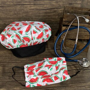 Può includere: Una maschera facciale in tessuto bianco e un cappello da cuoco in tessuto bianco con una fascia nera, entrambi con una stampa di anguria rossa. È presente anche uno stetoscopio blu.