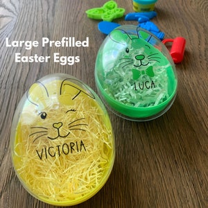 May include: Two large, clear plastic Easter eggs with bunny faces. The green egg has a white bow tie and the name "LUCA" written on it. The yellow egg has the name "VICTORIA" written on it.