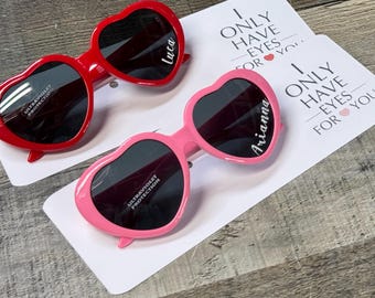 Könnte beinhalten: Zwei herzförmige Sonnenbrillen, eine rote und eine rosa, mit dunklen Gläsern. Das rote Paar hat den Namen "Luca" auf dem Glas, und das rosa Paar hat "Arianna". Beide Paare liegen auf weißen Karten mit dem Text "I ONLY HAVE EYES FOR YOU."
