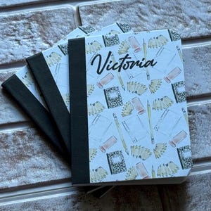 Könnte beinhalten: Mehrere Notizbücher mit weißem Einband, verziert mit Schulmaterialien und dem Namen "Victoria" in schwarzer Schrift. Die Notizbücher haben einen schwarzen Rücken und ein Muster.