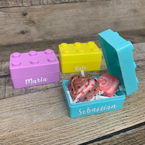 Könnte beinhalten: Drei kleine Plastikboxen in Form von Bausteinen, jede in einer anderen Farbe und mit einem Namen auf der Vorderseite. Die Boxen sind mit kleinen Schokoladen gefüllt. Die Namen auf den Boxen sind Maria, Kate und Sebastian.