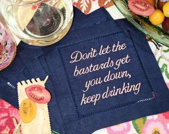 Puede incluir: Posavasos de lino azul marino con texto bordado: "Don't let the bastards get you down, keep drinking." Se ven una copa de vino blanco y una botella de sidra rosada. La escena incluye un plato de tomates coloridos.