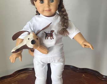 May include: A doll wearing a white t-shirt with an embroidered dog design and white pants. The doll is holding a brown and white plush dog.