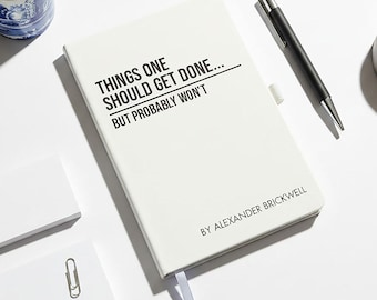 May include: A white notebook with the text "THINGS ONE SHOULD GET DONE... BUT PROBABLY WON'T" and the author's name. A black pen, a small gray clock, a blue and white patterned mug with coffee, and two white notepads with a paperclip are also present.