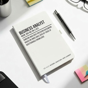 May include: A white notebook with the text "BUSINESS ANALYST" and a humorous definition. The image also includes a cup of coffee, glasses, pens, and sticky notes, suggesting an office or workspace setting.
